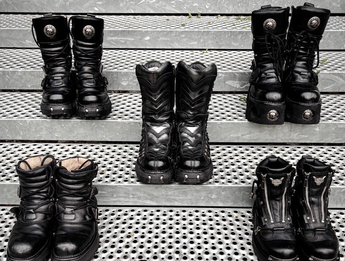 Six pairs of black fashion footwear arranged on metal perforated steps: four tall black boots and two black lace-up shoes, photographed from above.