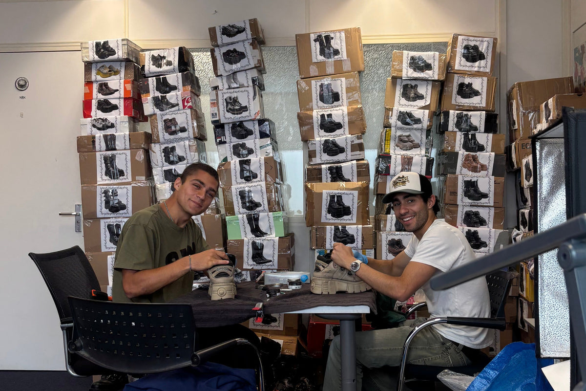 A workshop scene with two men seated at a table working on what appear to be shoe-related projects, with shelves and boxes of shoe boxes behind them. The foreground is cluttered with tools, a blue spray can, a red container, and other workshop items.