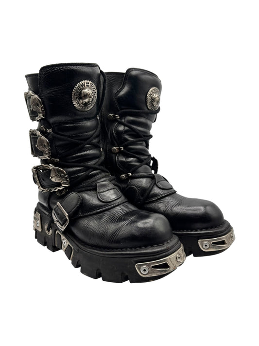 Black mid-calf lace-up boots with straps, buckles, and raised metal guard plates
