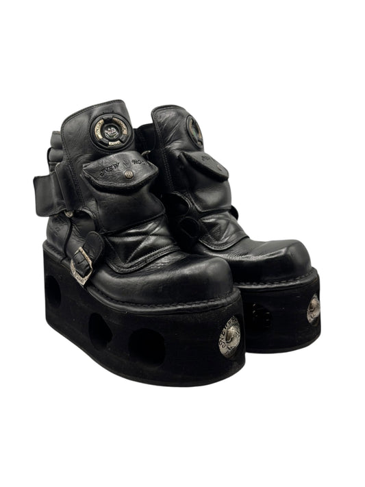 Black platform boots, side view with circular logo patch, straps, and thick lug sole.