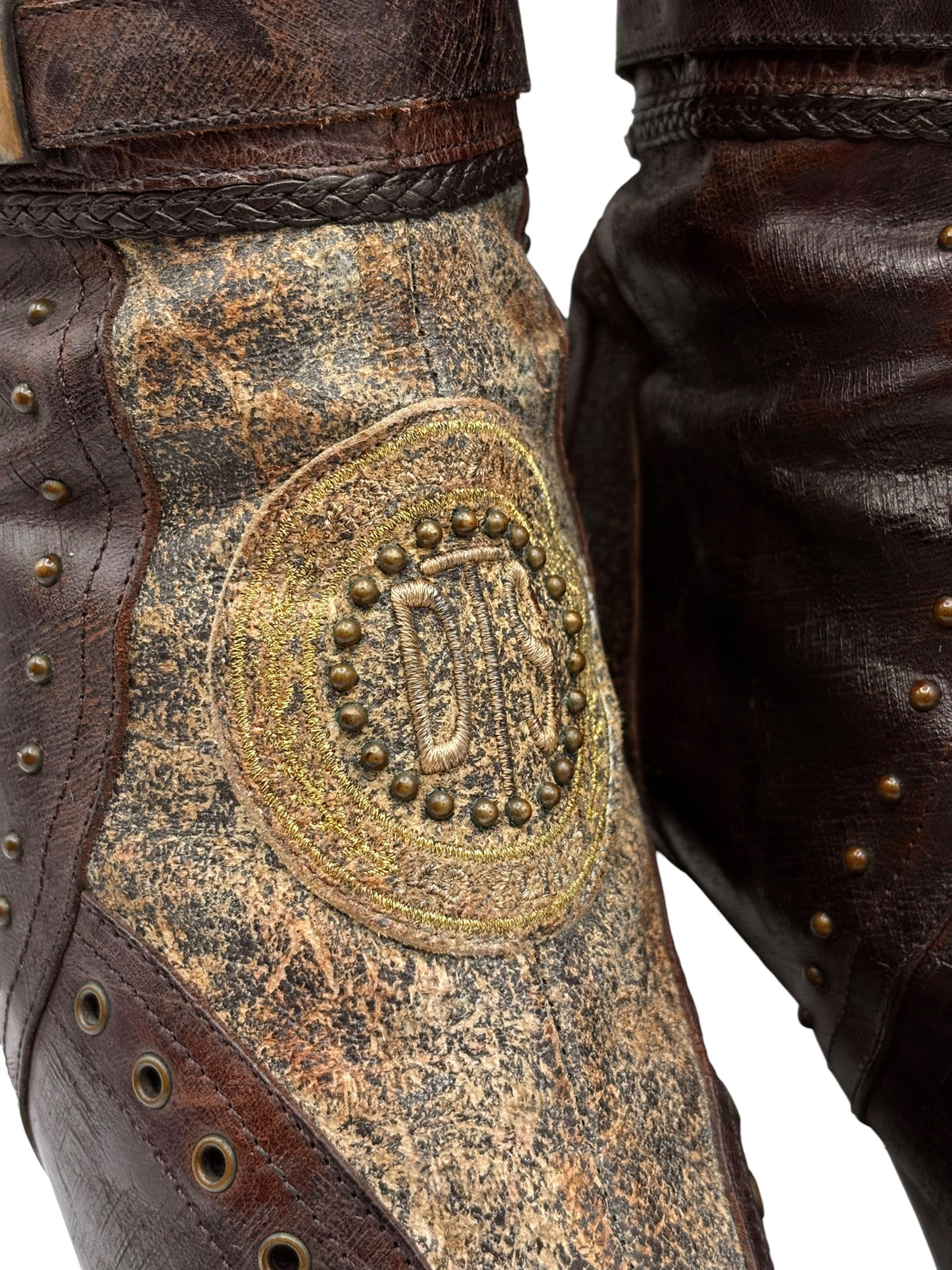 Knee-high boots detail: round gold emblem on distressed toe leather with studded trim