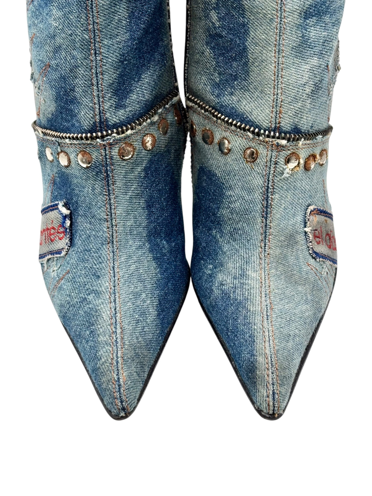 Pair of mid-calf denim boots with pointed toes, rivet trim, and decorative patches